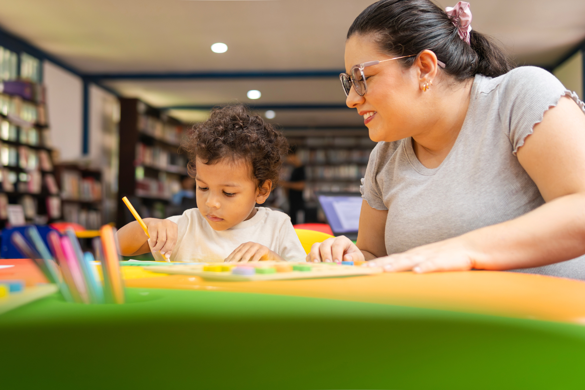 Latina teacher smiling as she guides her preschool student in an art project where the child is coloring. A library is visible in the background