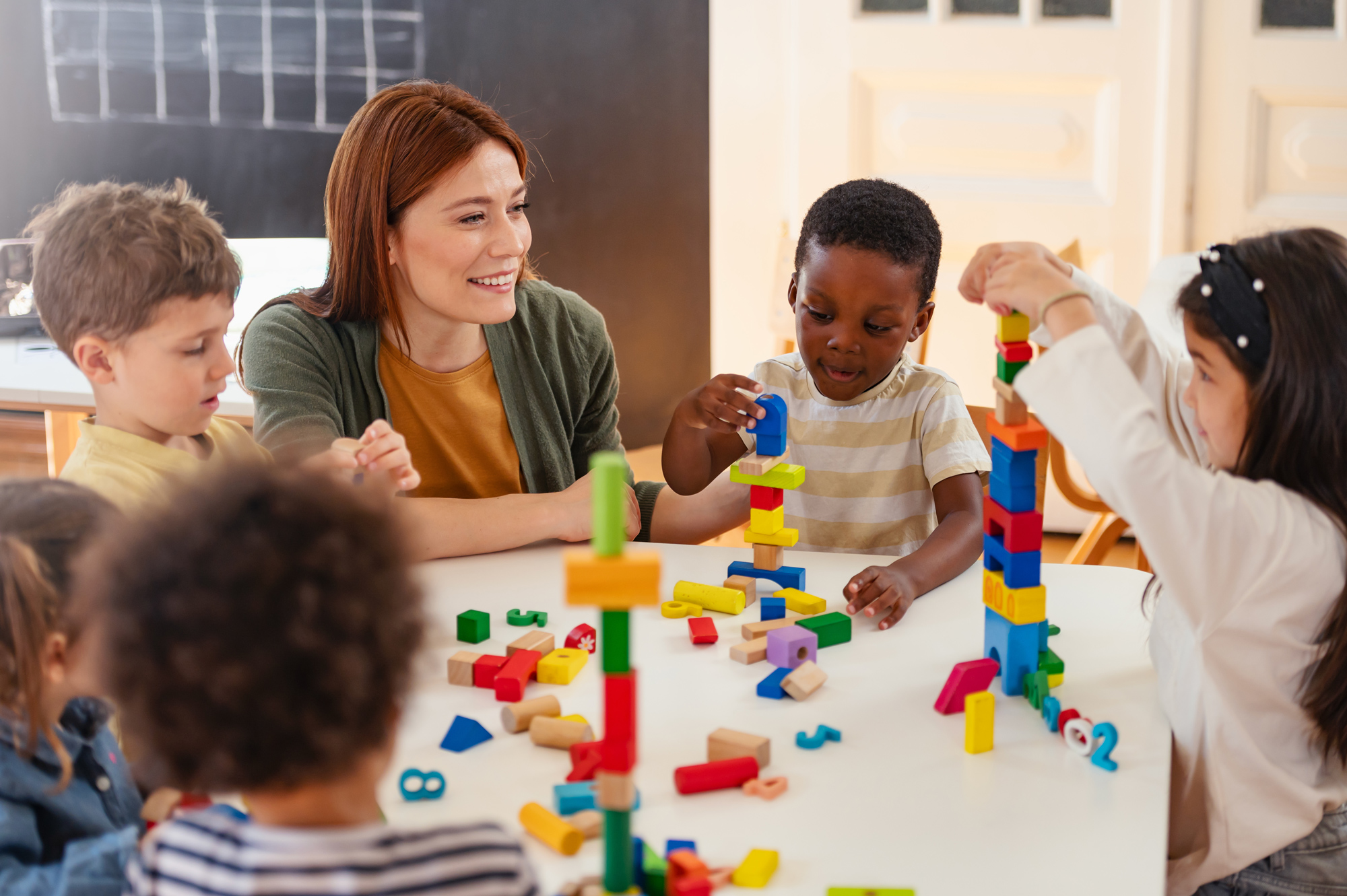 Diverse preschool children play with colorful building blocks at a classroom table, guided by a smiling teacher in a creative, inclusive early learning environment